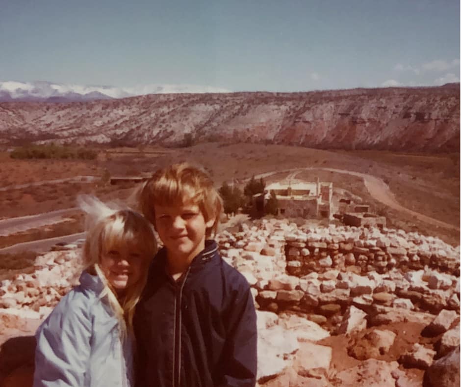 Two young kids about the age of six and eight stand in front of hills in the background and stone structures in the foreground. The stones make up the shape of walls and other structures. 