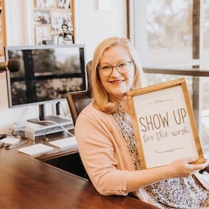 Woman sits at a desk, holding a sign that reads: "Show up for the work."