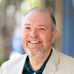 Man with a beard and short hair wearing a tan blazer smiles at the camera